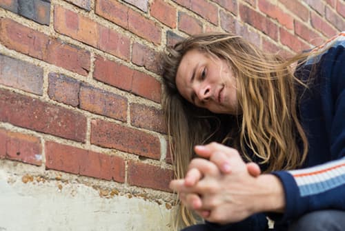 Man portraying an addict leaning against brick wall outside